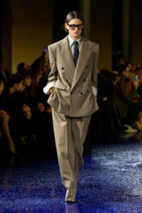 Model on a fashion runway wearing a large beige double-breasted suit with a blue tie and sunglasses, hands in pockets.