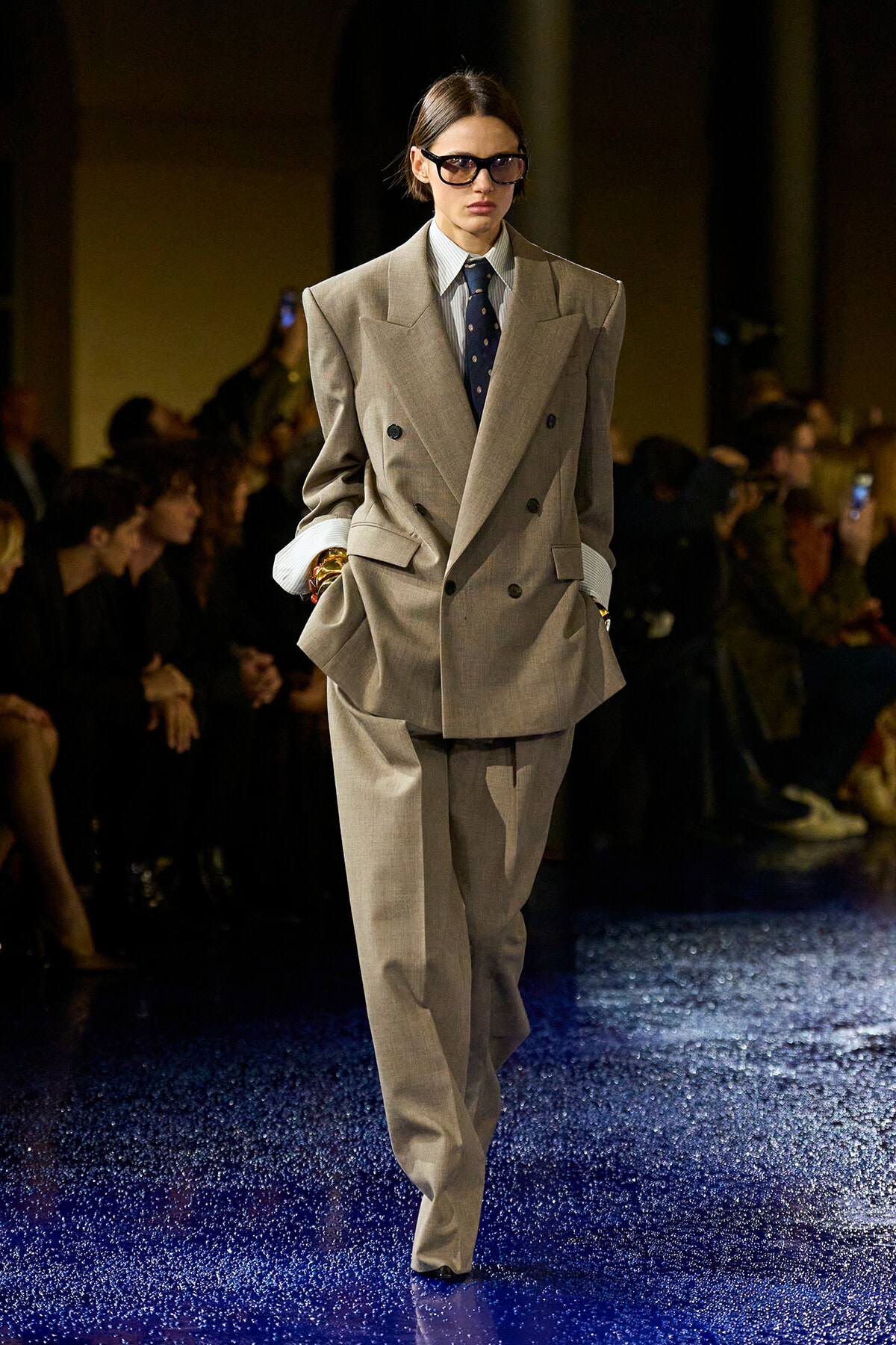 Model on a fashion runway wearing a large beige double-breasted suit with a blue tie and sunglasses, hands in pockets.