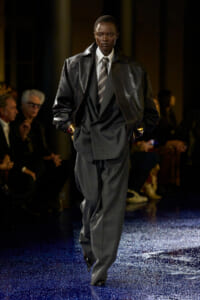 Male model in an oversized black leather jacket over a gray suit and striped tie walks a blue-glitter runway, hands in pockets.