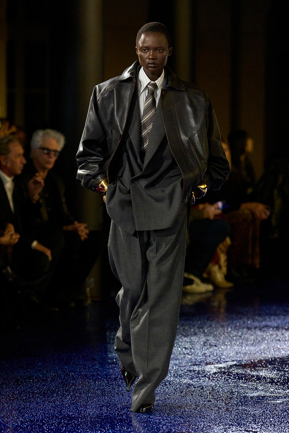 Male model in an oversized black leather jacket over a gray suit and striped tie walks a blue-glitter runway, hands in pockets.
