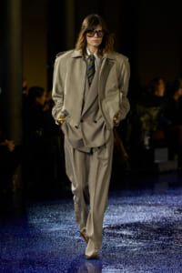Model on a runway wearing a loose beige suit with a tie, oversized jacket, and sunglasses under spotlight.