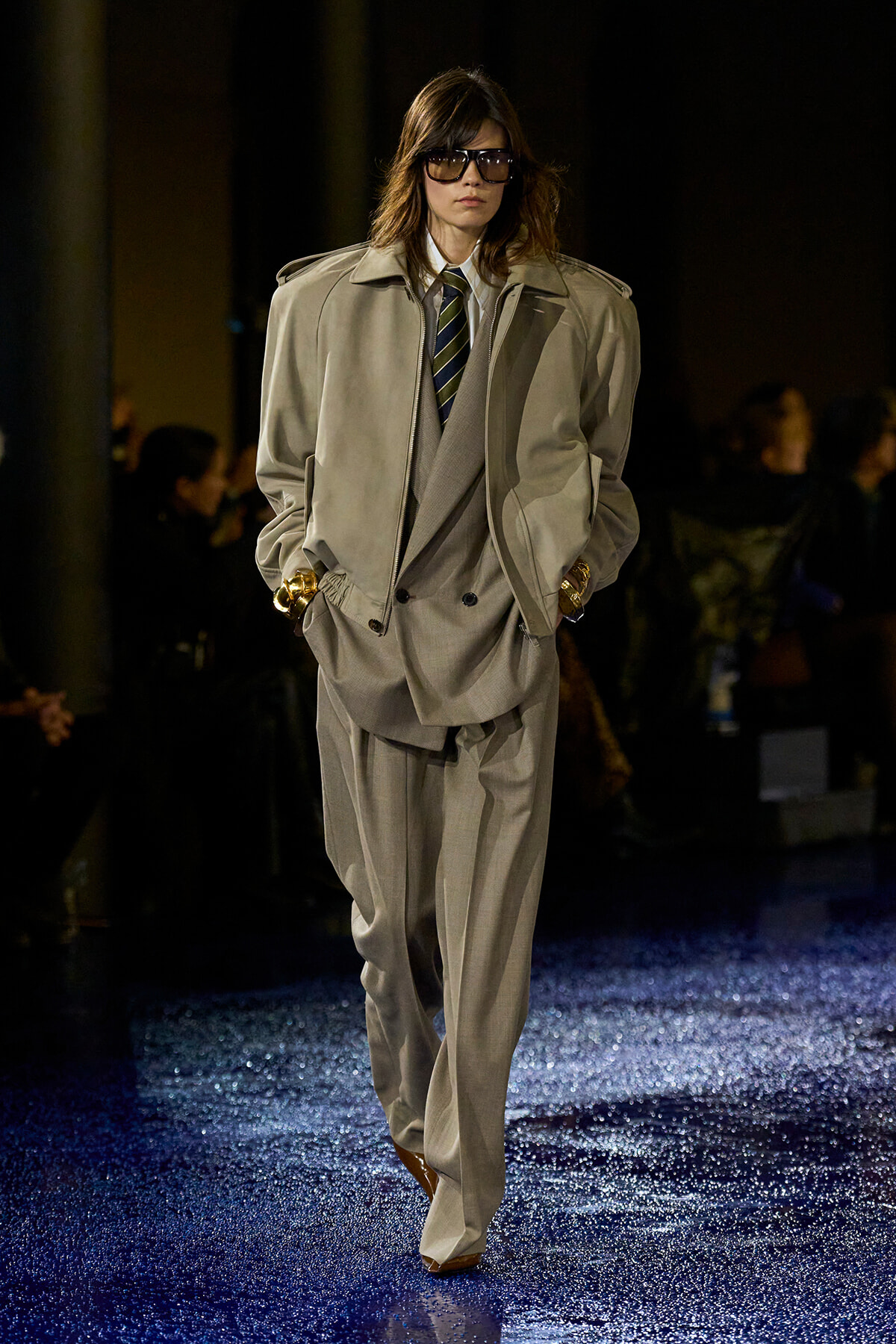 Model on a runway wearing a loose beige suit with a tie, oversized jacket, and sunglasses under spotlight.