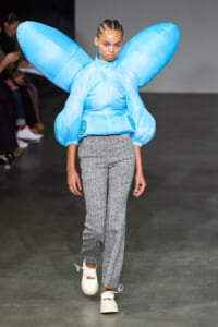 Model walking a runway in a bright blue oversized puffer with wing-like sleeves, paired with gray textured pants and white sneakers.