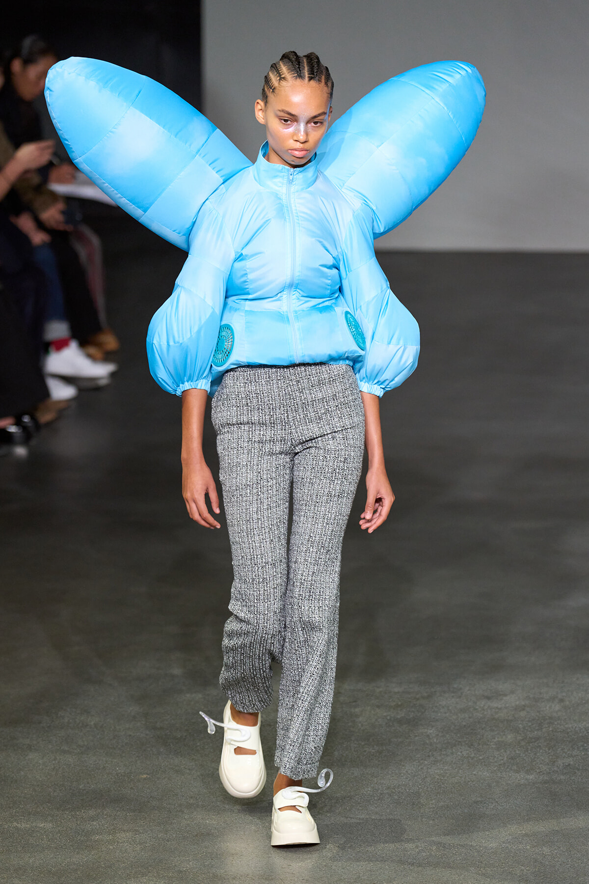 Model walking a runway in a bright blue oversized puffer with wing-like sleeves, paired with gray textured pants and white sneakers.