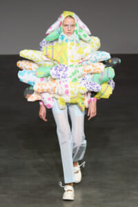 Model on a runway wearing an oversized multicolor puffer with balloon-like panels around the torso and a hood, forming a flower/bouquet silhouette.