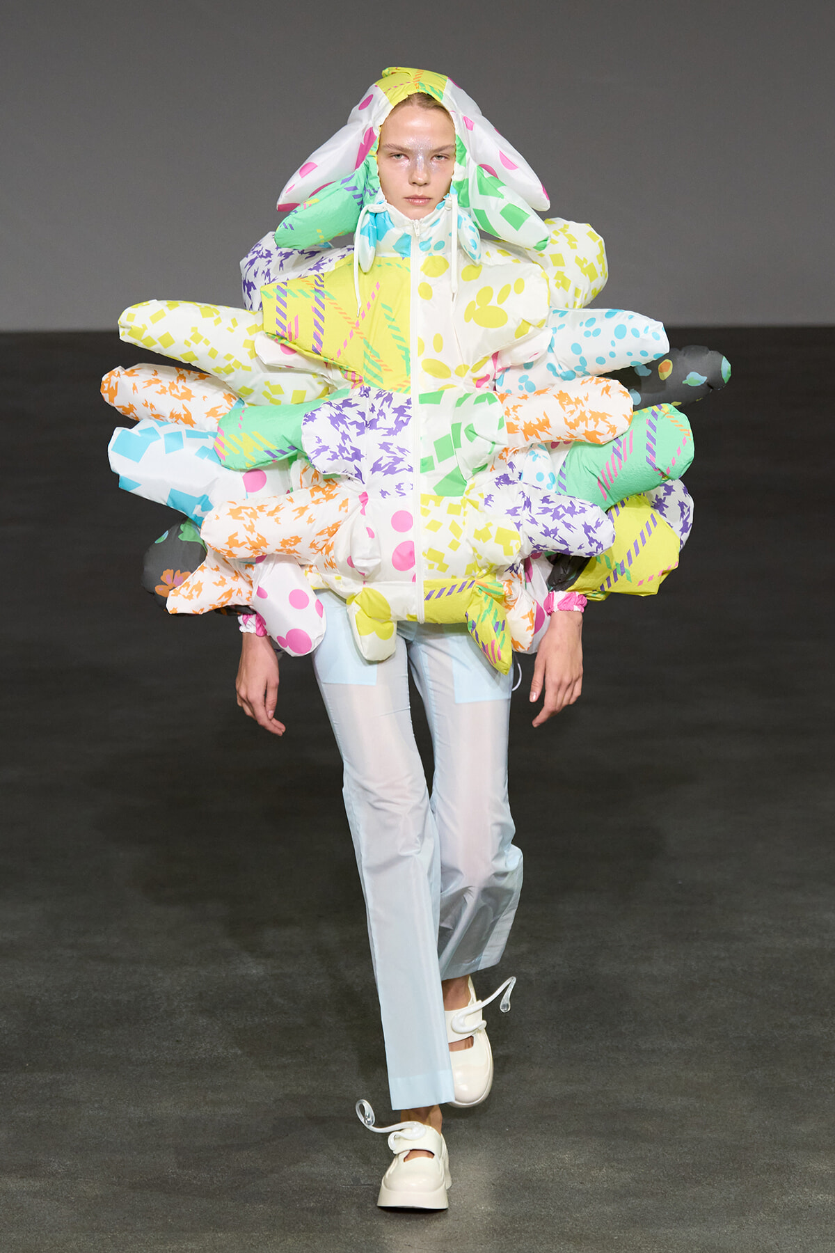 Model on a runway wearing an oversized multicolor puffer with balloon-like panels around the torso and a hood, forming a flower/bouquet silhouette.