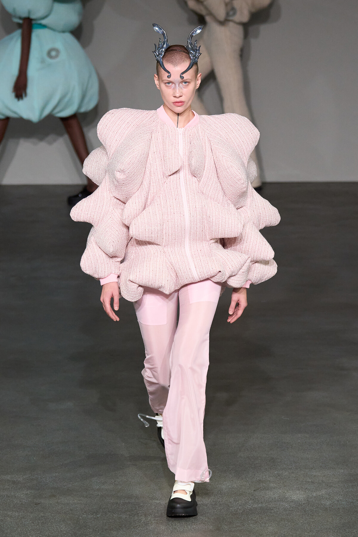 Model on runway wearing an oversized pink textured sweater with large ruffled sculpted sleeves and pale pink trousers, plus metallic horn headpiece.
