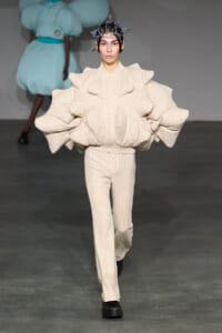 Model walks the runway in a cream textured suit with oversized sculptural sleeves and a metallic headpiece.