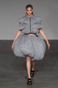 Runway model wearing a chunky, textured gray two-piece outfit with a voluminous bubble skirt and oversized puffed sleeves, on a gray backdrop.