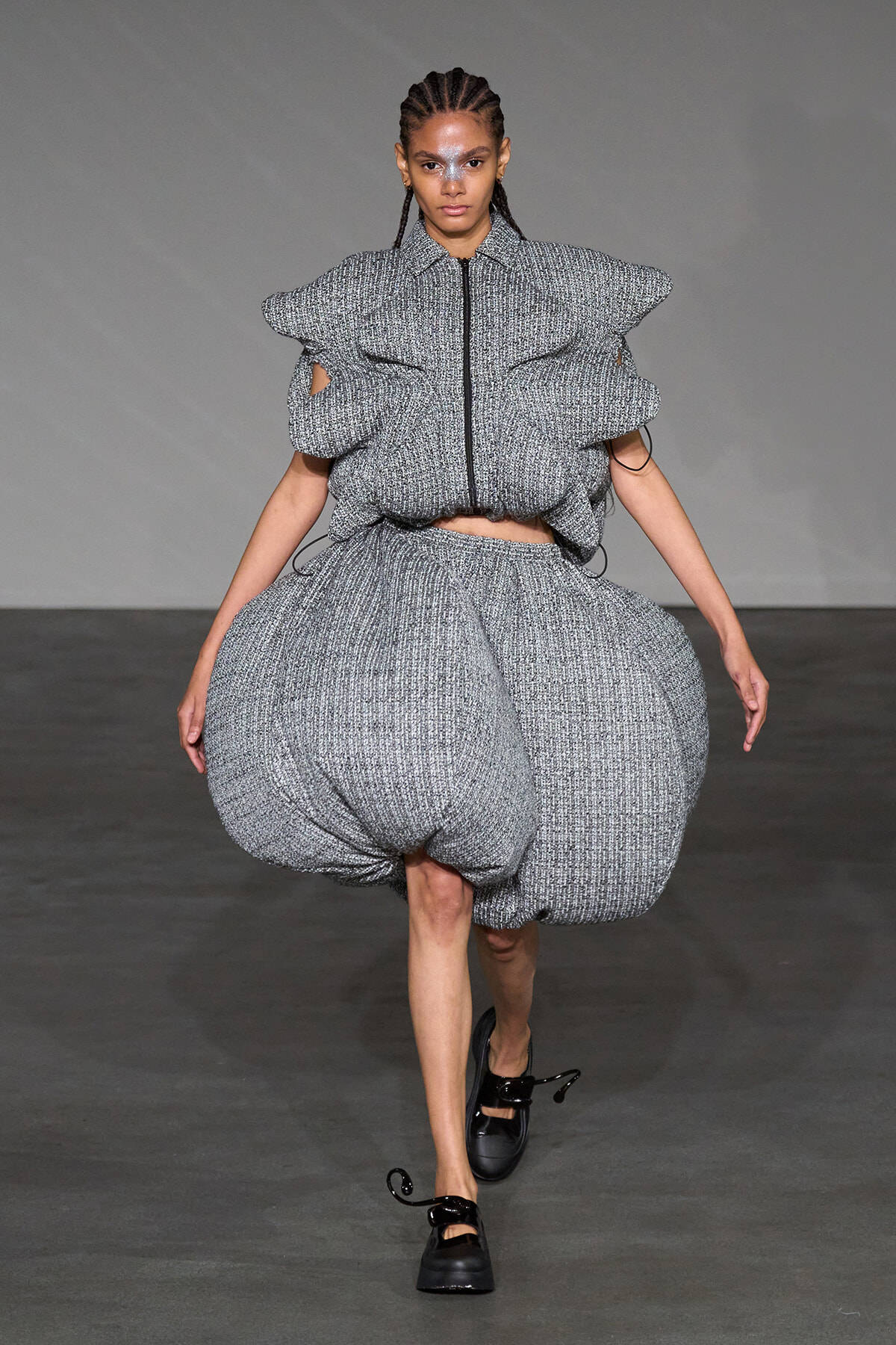 Runway model wearing a chunky, textured gray two-piece outfit with a voluminous bubble skirt and oversized puffed sleeves, on a gray backdrop.
