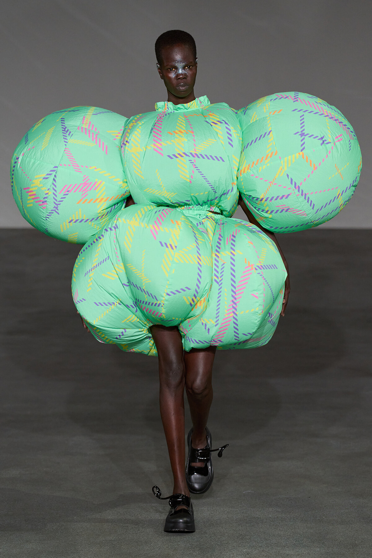 Model walking a runway in a mint-green inflatable outfit with oversized circular shoulders and hips, featuring purple and yellow striped patterns.