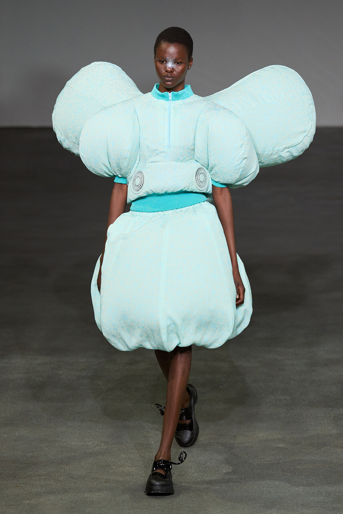 Model wearing a futuristic light-blue sculptural outfit with oversized shoulder puffs and a bubble skirt on a runway.