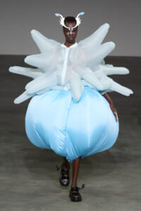 Model on runway wearing an oversized pale blue inflated skirt with white sculptural top and antler headpiece, black shoes visible.