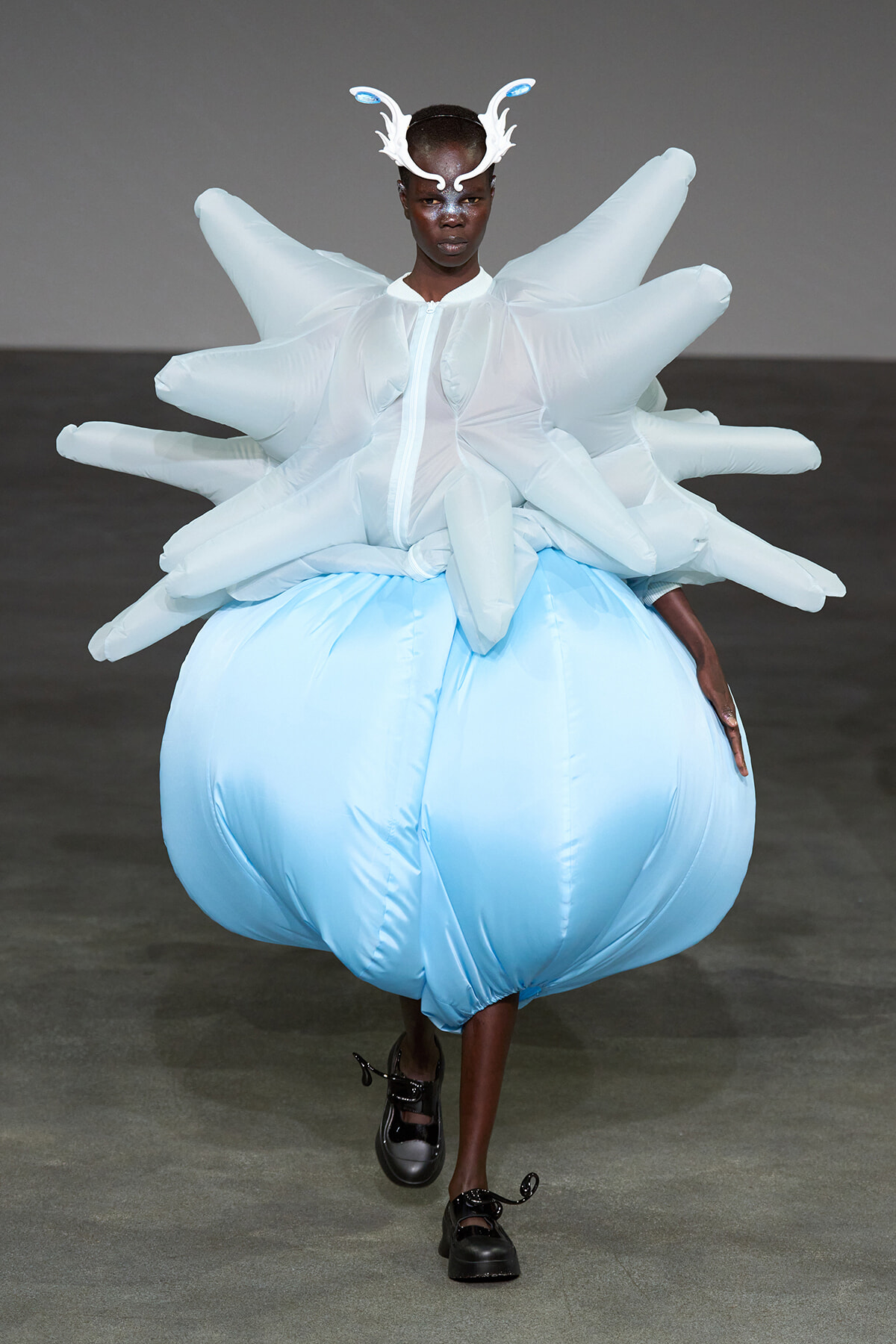 Model on runway wearing an oversized pale blue inflated skirt with white sculptural top and antler headpiece, black shoes visible.