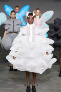 Model wearing a voluminous white cloud-like dress with a high collar and antler-inspired headpiece on a fashion runway