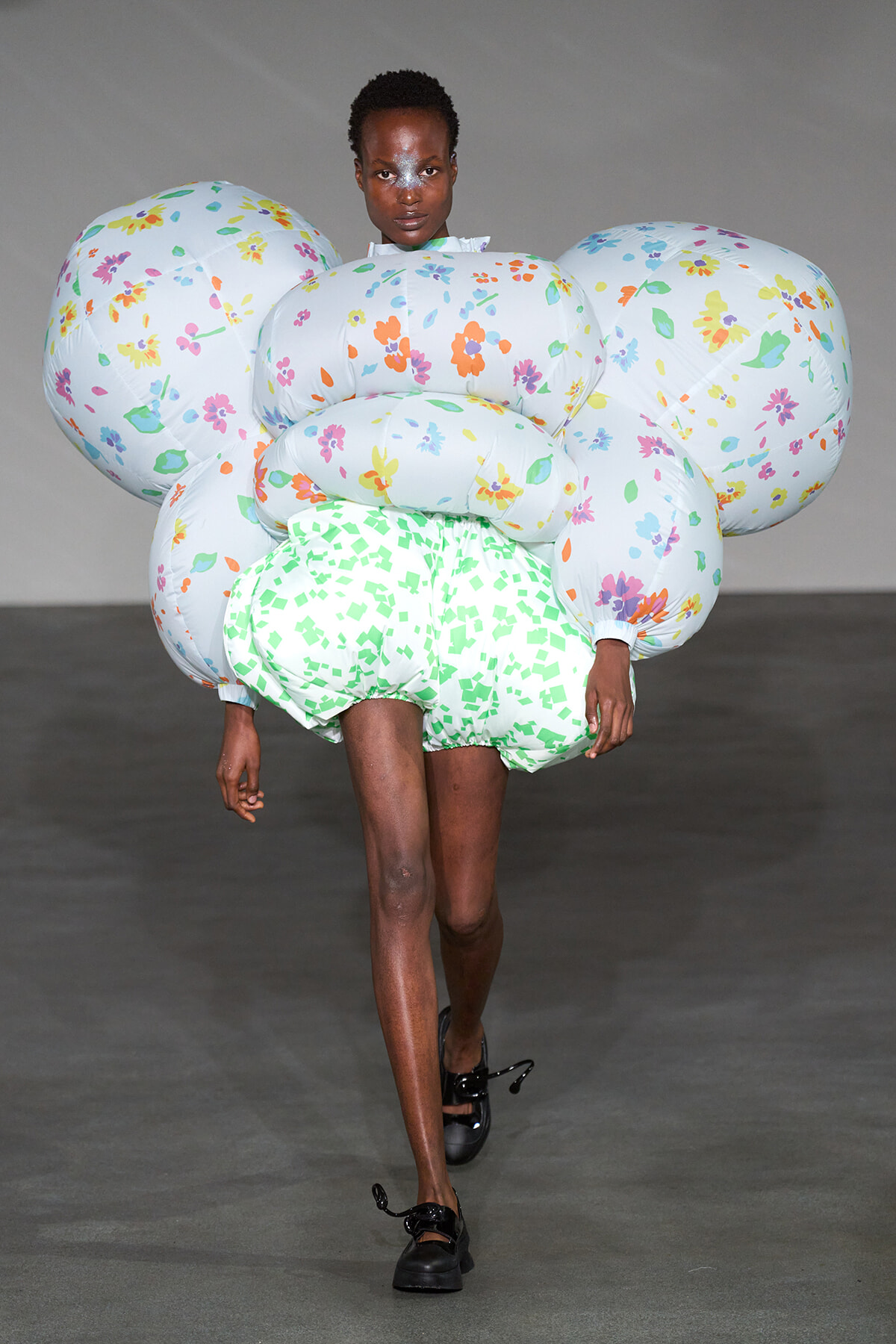 Runway model wearing a large, inflatable floral dress with multiple rounded, balloon-like segments in pastel blue with colorful flowers and a green speckled bottom.