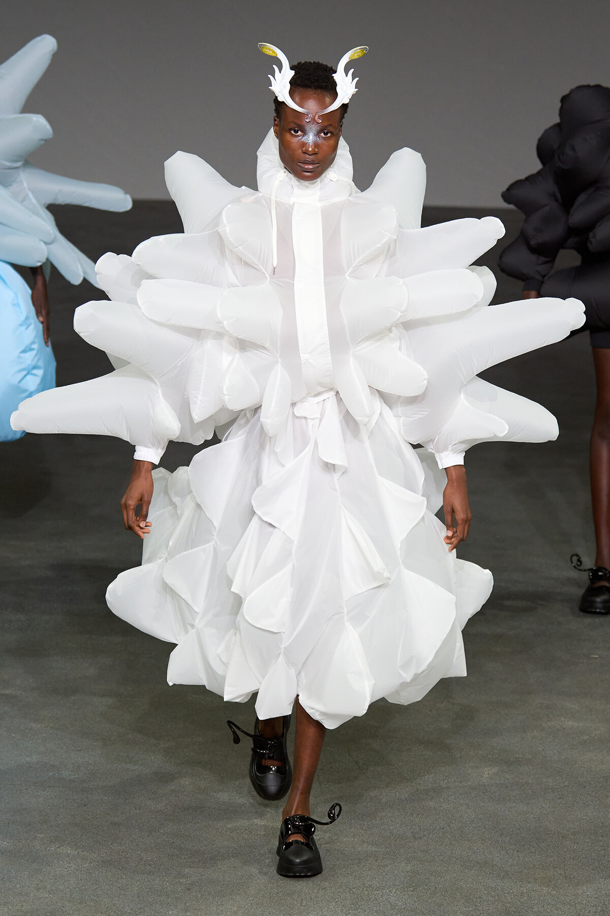 Model walks the runway in a white, sculptural gown with voluminous, inflatable petals and a horned headpiece resembling antlers.