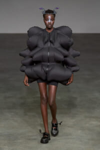 Model walking the runway in a dramatic oversized black inflatable suit with voluminous sculpted shoulders, paired with black shoes and a headpiece.
