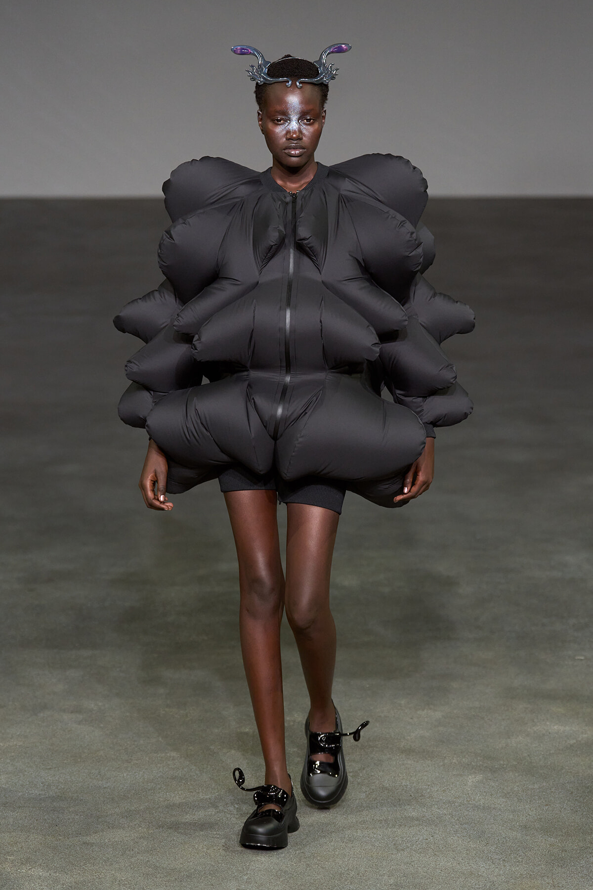 Model walking the runway in a dramatic oversized black inflatable suit with voluminous sculpted shoulders, paired with black shoes and a headpiece.