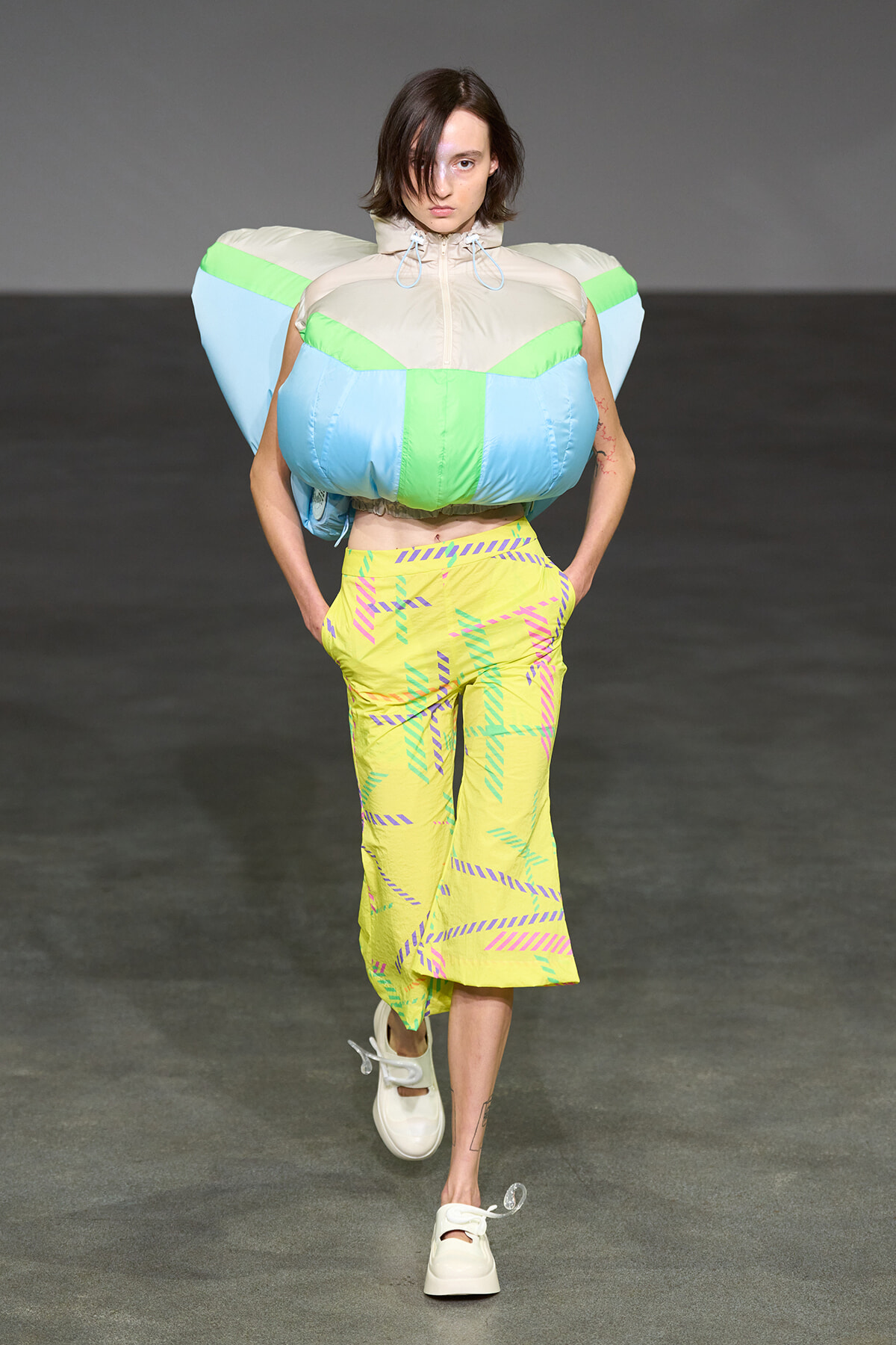 Model walking the runway in a neon color-blocked outfit with oversized capsule-shaped shoulders and pastel-yellow pants with pink and teal patterns, white sneakers.