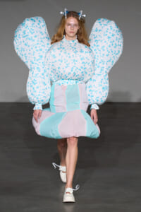 Model walking a runway in a pastel geometric dress with oversized bubble sleeves and light blue-pink panels, wearing a butterfly-like headpiece.