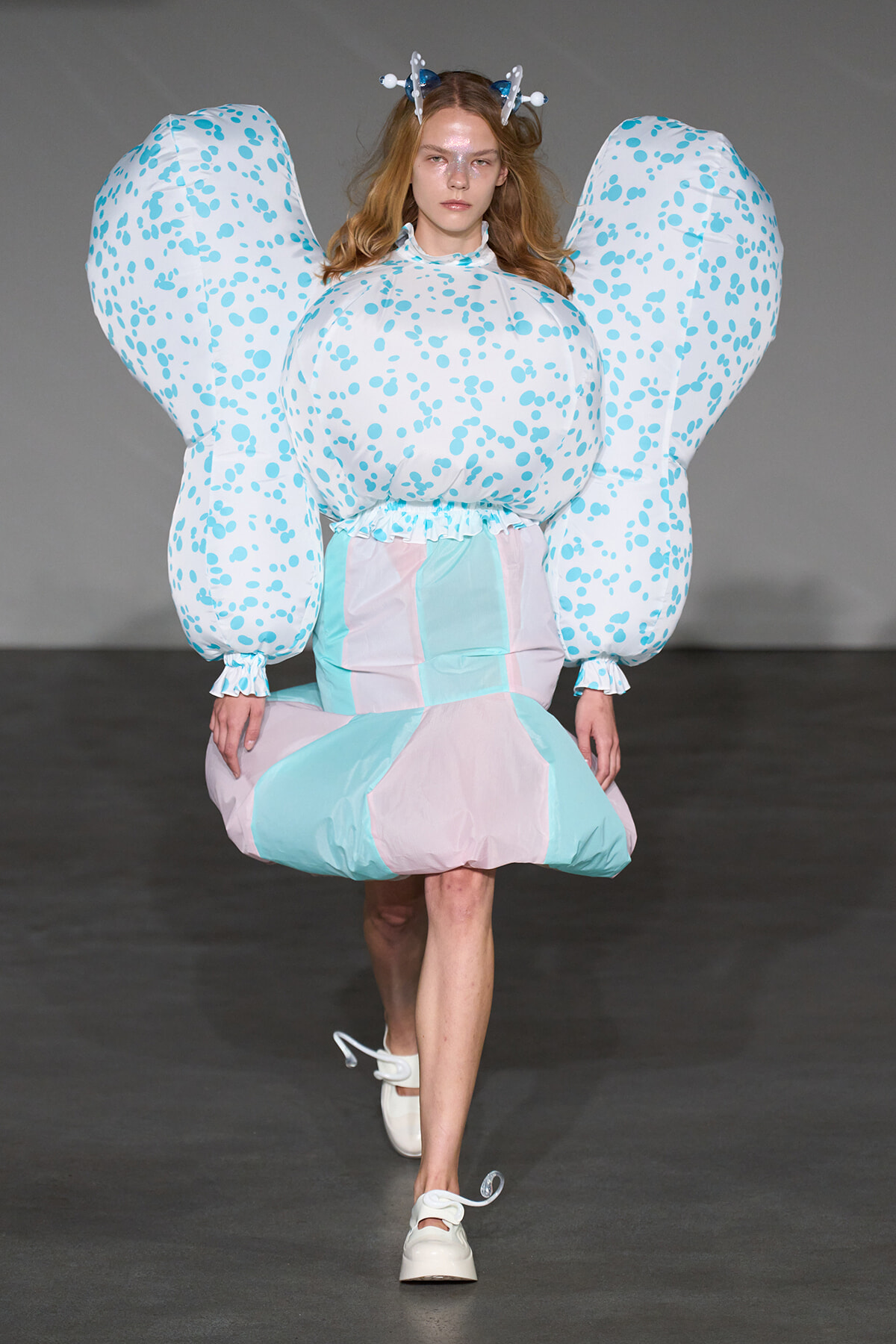 Model walking a runway in a pastel geometric dress with oversized bubble sleeves and light blue-pink panels, wearing a butterfly-like headpiece.