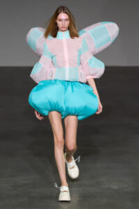 Model on runway wearing an oversized pastel pink and turquoise padded jacket with large wing-like sleeves and turquoise bubble shorts.