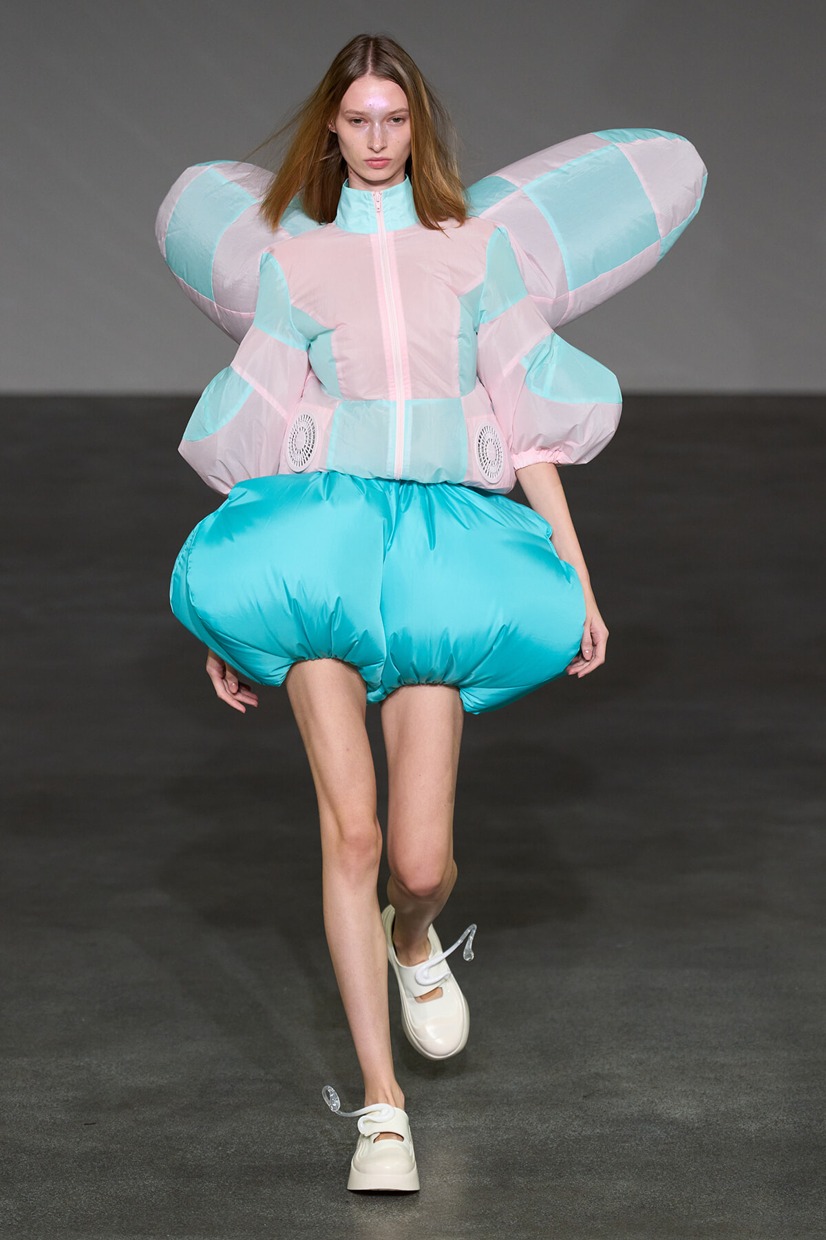 Model on runway wearing an oversized pastel pink and turquoise padded jacket with large wing-like sleeves and turquoise bubble shorts.