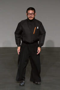 Male model on a gray runway wearing an all-black outfit: a puffer jacket with an orange zipper accent and wide-leg pants, black sneakers, and round glasses.