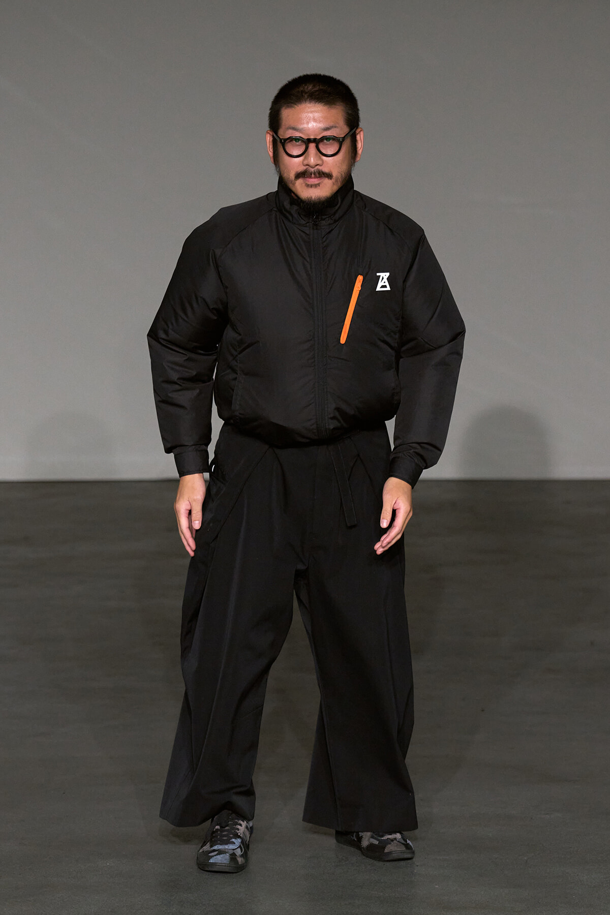 Male model on a gray runway wearing an all-black outfit: a puffer jacket with an orange zipper accent and wide-leg pants, black sneakers, and round glasses.
