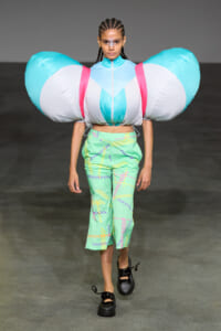 Model walks the runway in an oversized inflatable shoulder piece in turquoise, white, and pink, paired with mint green patterned pants and black shoes.