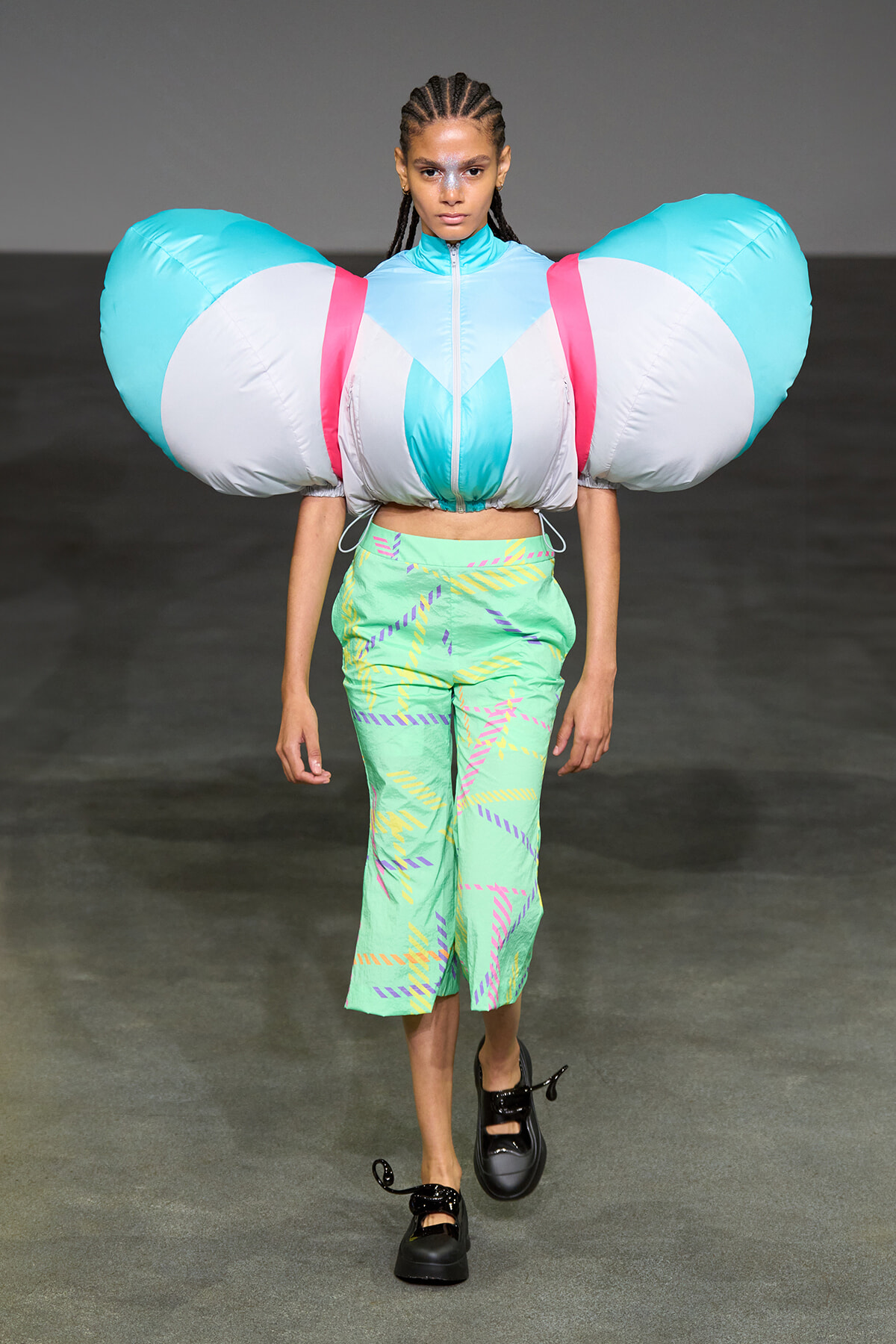 Model walks the runway in an oversized inflatable shoulder piece in turquoise, white, and pink, paired with mint green patterned pants and black shoes.