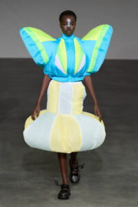 Model on a runway wearing a futuristic dress with oversized blue and neon-green wing sleeves and a pale-yellow items skirt, dramatic and sculptural.