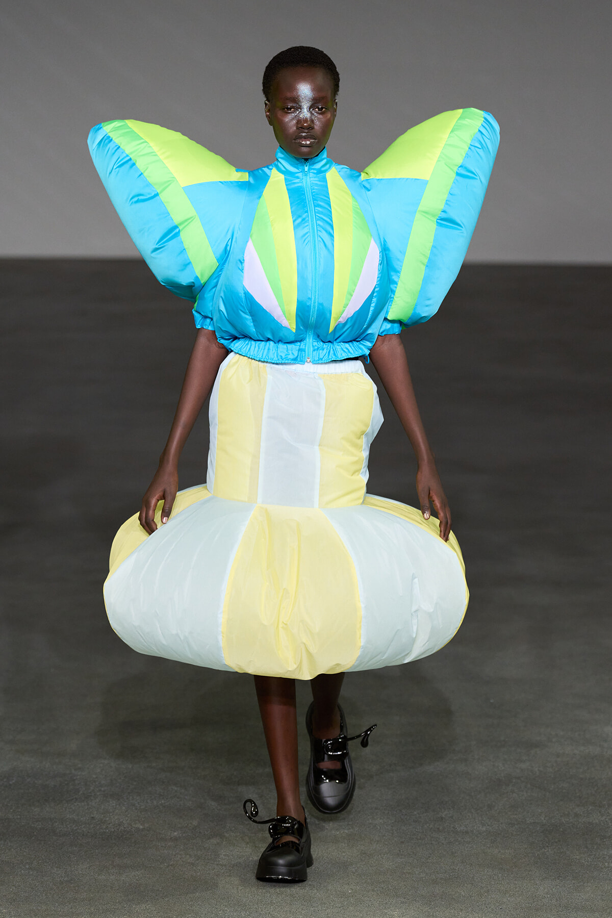 Model on a runway wearing a futuristic dress with oversized blue and neon-green wing sleeves and a pale-yellow items skirt, dramatic and sculptural.