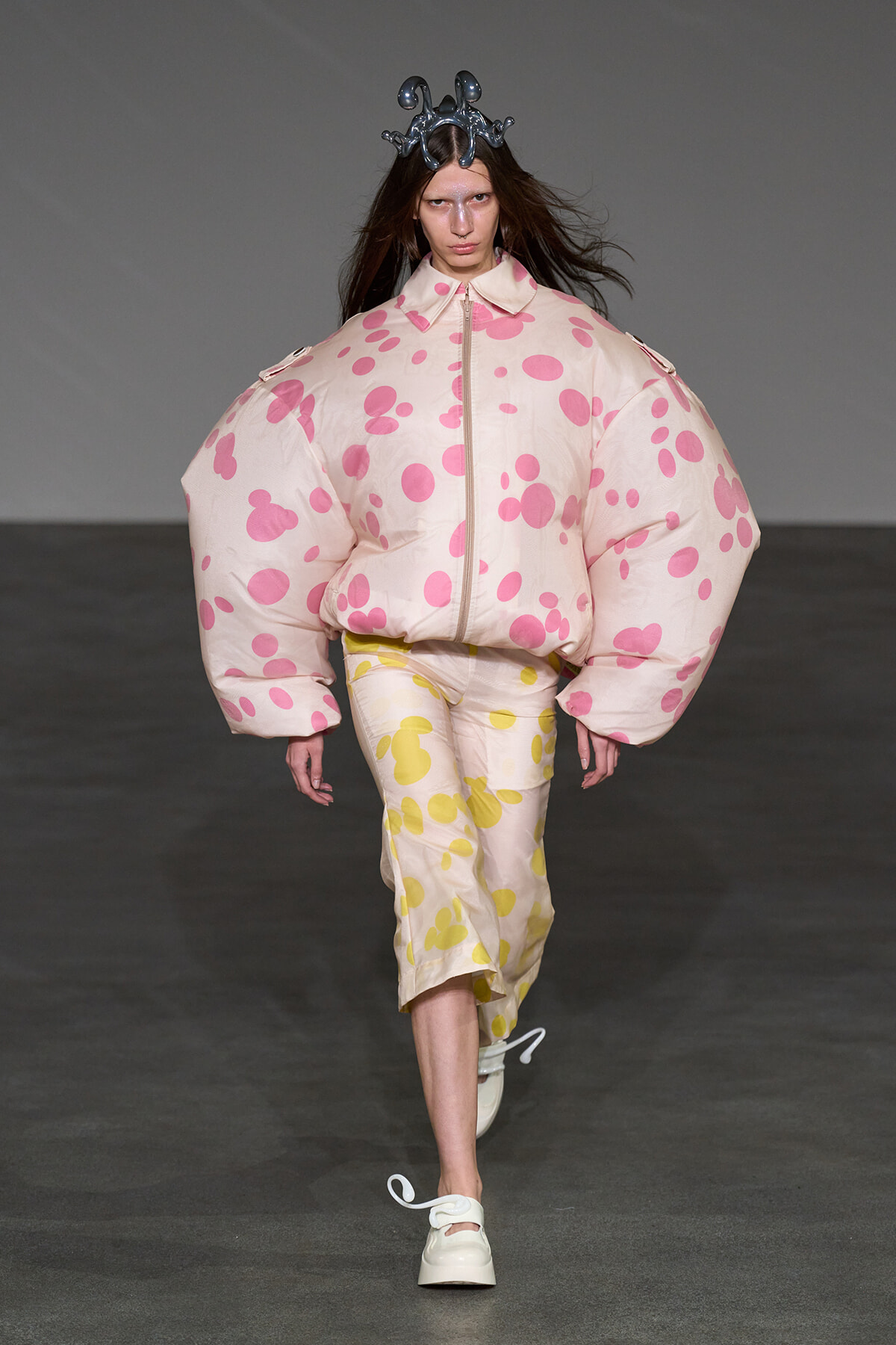 Model walks the runway in a voluminous pale pink polka-dot puffer jacket with matching pants and a metallic crown headpiece.