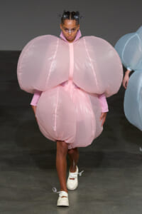 Model walking the runway in a voluminous pastel pink bubble dress with oversized sleeves and structured shoulders, paired with white sneakers and futuristic hair accessories.