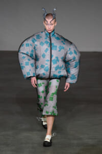 Model walking runway in a silver oversized puffer with teal square patterns and matching capri pants, wearing a metallic horned headpiece.