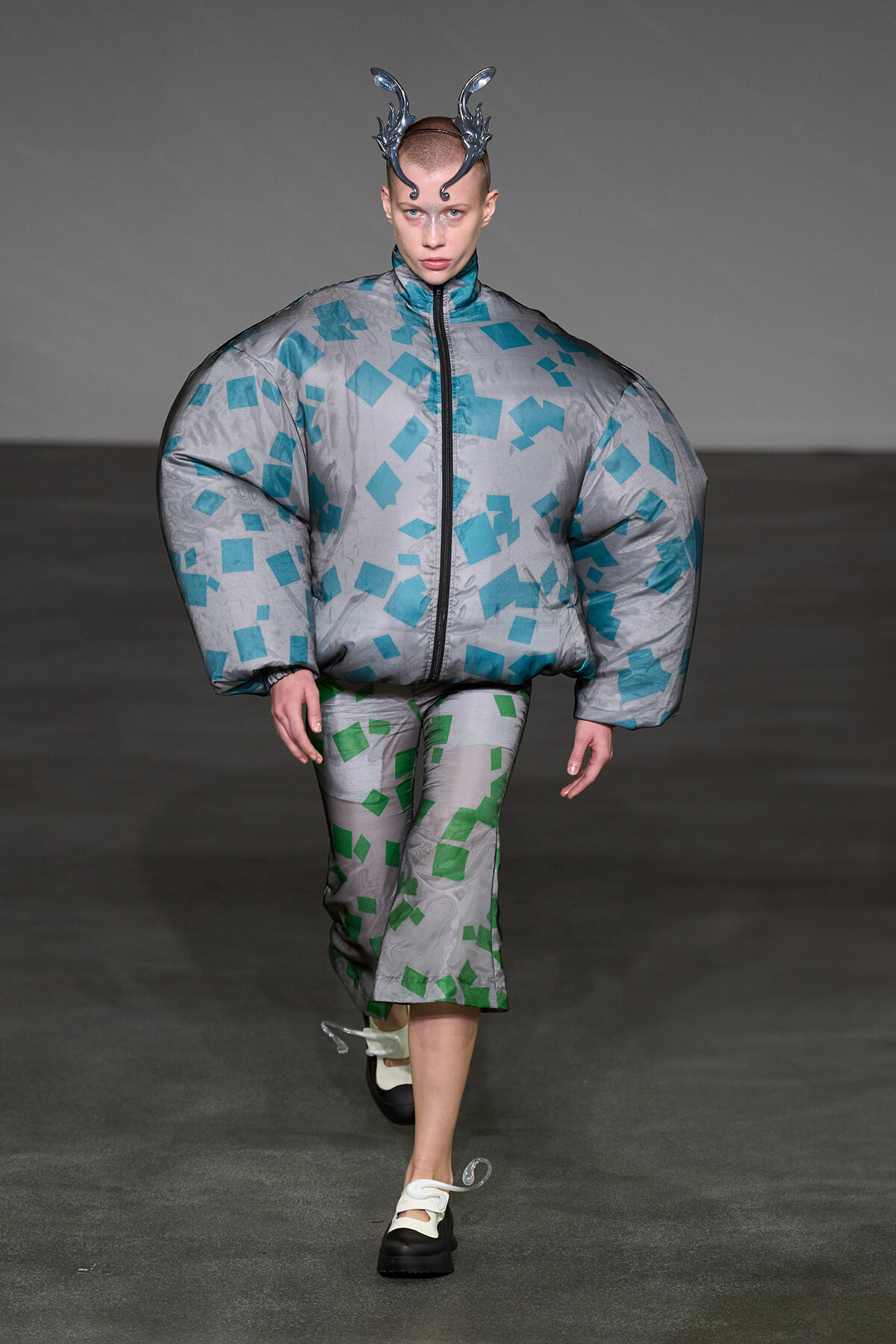 Model walking runway in a silver oversized puffer with teal square patterns and matching capri pants, wearing a metallic horned headpiece.