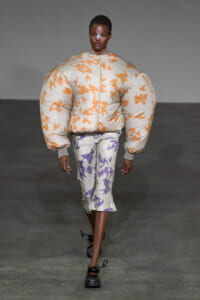Model walking a runway in a light gray oversized puffer jacket with orange pattern and matching oversized pants in lavender accents