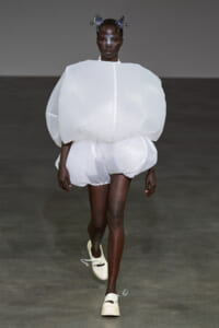 Model walks a runway in an oversized, white sculptural ensemble with a large rounded torso and bubble-like shorts, paired with pale shoes and a metallic headpiece.