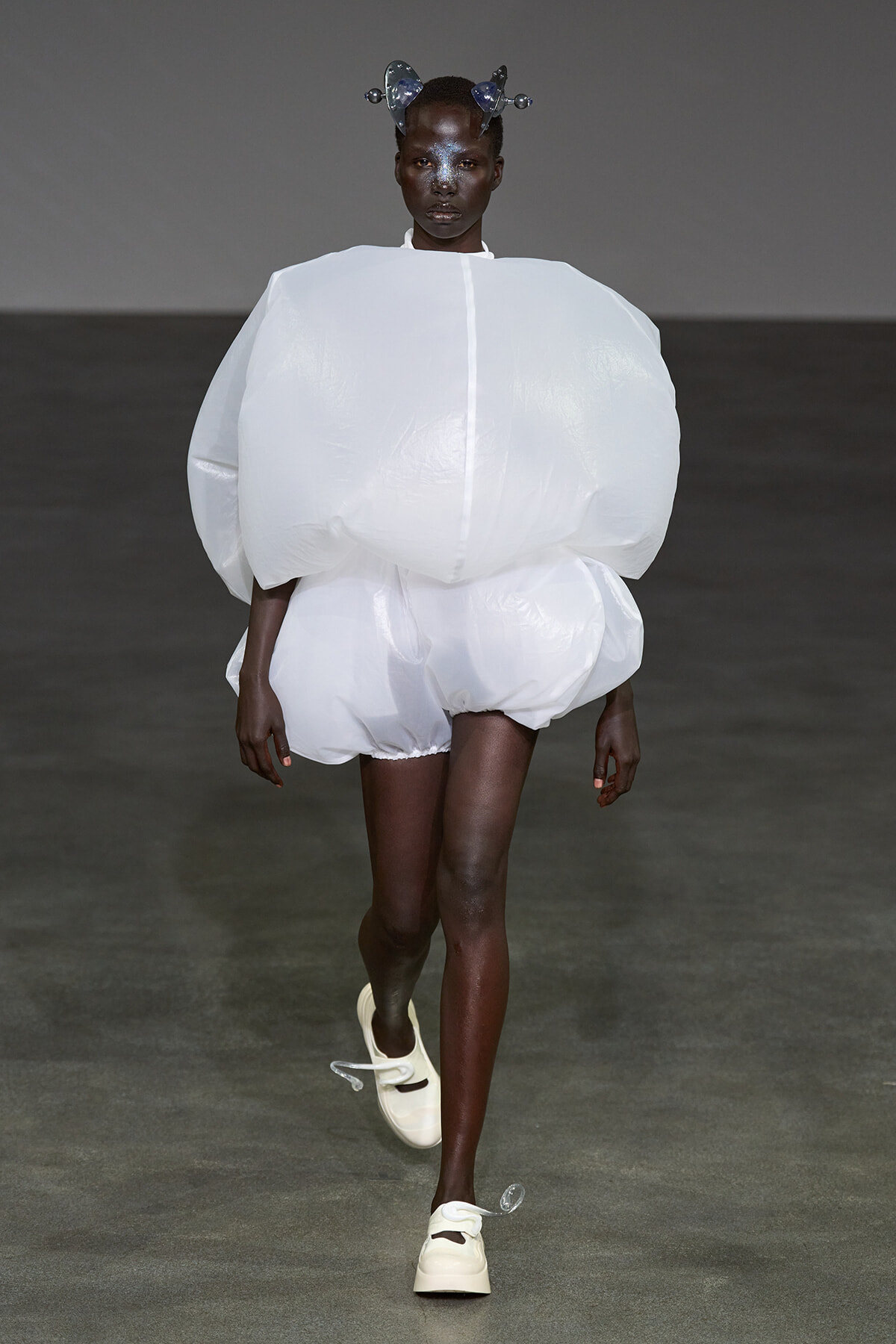 Model walks a runway in an oversized, white sculptural ensemble with a large rounded torso and bubble-like shorts, paired with pale shoes and a metallic headpiece.
