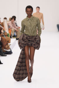 Model walks a fashion runway in an olive-green plaid button-down shirt with a large draped plaid skirt and brown heels.