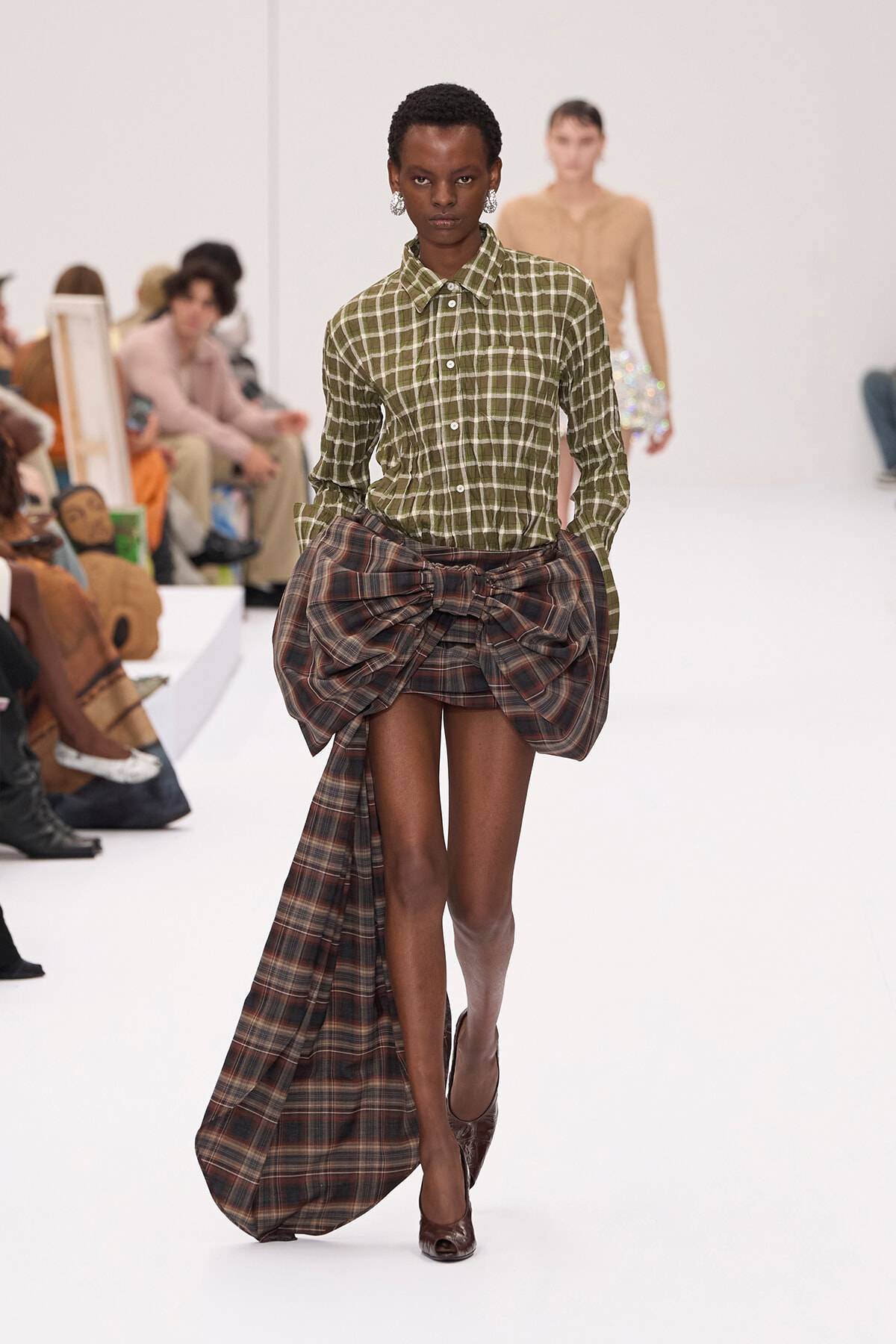 Model walks a fashion runway in an olive-green plaid button-down shirt with a large draped plaid skirt and brown heels.