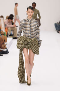 Model walking a white runway in a brown plaid button-down shirt and a large green plaid bow at the waist, paired with olive heels.