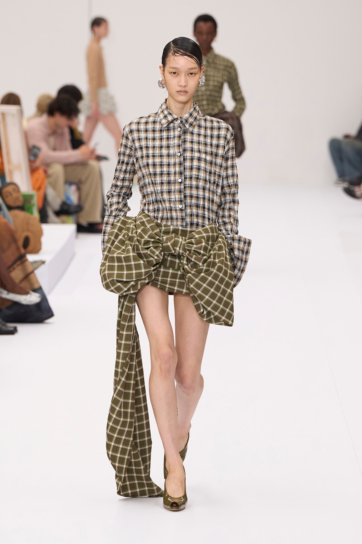Model walking a white runway in a brown plaid button-down shirt and a large green plaid bow at the waist, paired with olive heels.