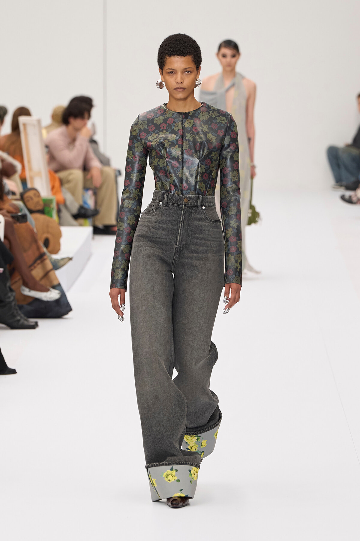 Front-on view of a fashion model walking a white runway in a dark floral long-sleeve top tucked into high-waisted gray jeans with oversized floral platform shoes; audience seated along the sides in the background.