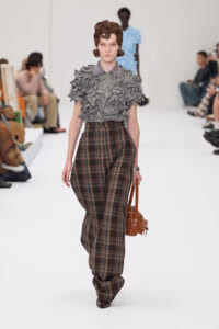 Model walks a runway wearing a gray ruffled short-sleeve top and high-waisted plaid wide-leg pants, carrying a brown handbag.