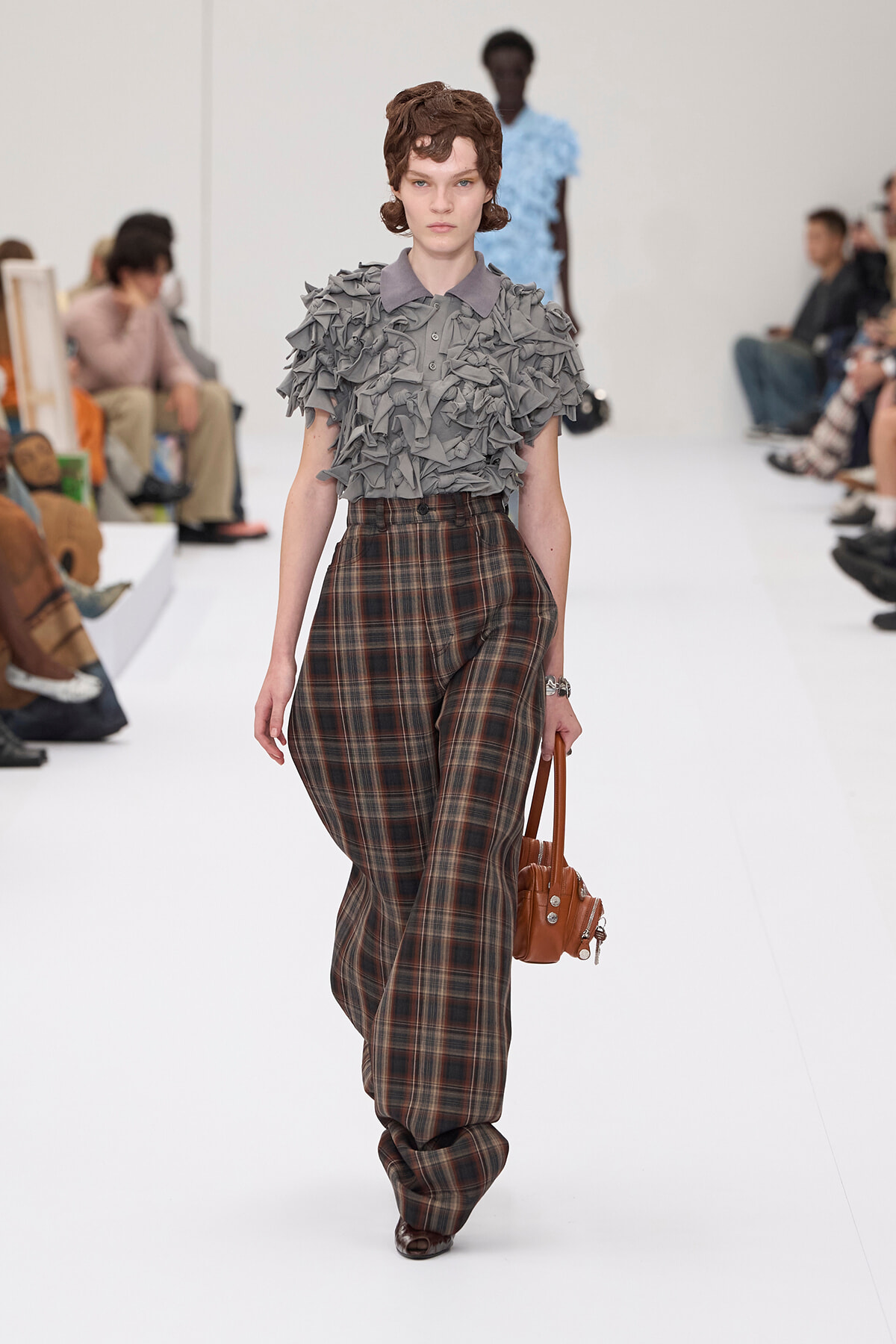 Model walks a runway wearing a gray ruffled short-sleeve top and high-waisted plaid wide-leg pants, carrying a brown handbag.