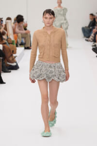 Model walks a runway wearing a beige cropped cardigan and a silver textured ruffled mini skirt, mint-green knit flats.