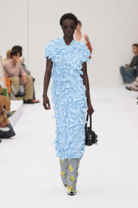 Model on a runway wearing a pale blue, petal-textured dress over floral-print pants, carrying a black handbag.